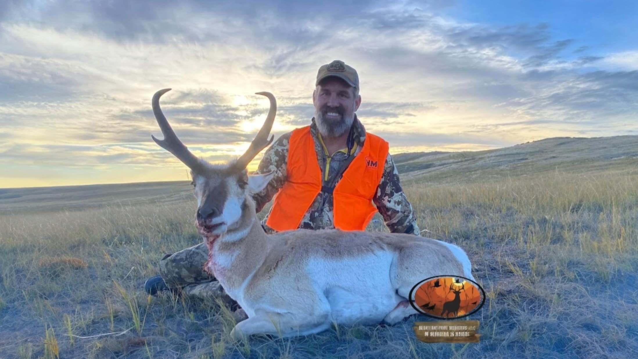 Hunter poses with a pronghorn antelope in a field. Antelope has large horns, white belly, and blood around the mouth. Hunt...