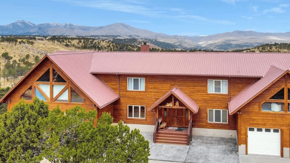 Large wooden lodge with a red metal roof. Mountains in the background. Green trees in the front.