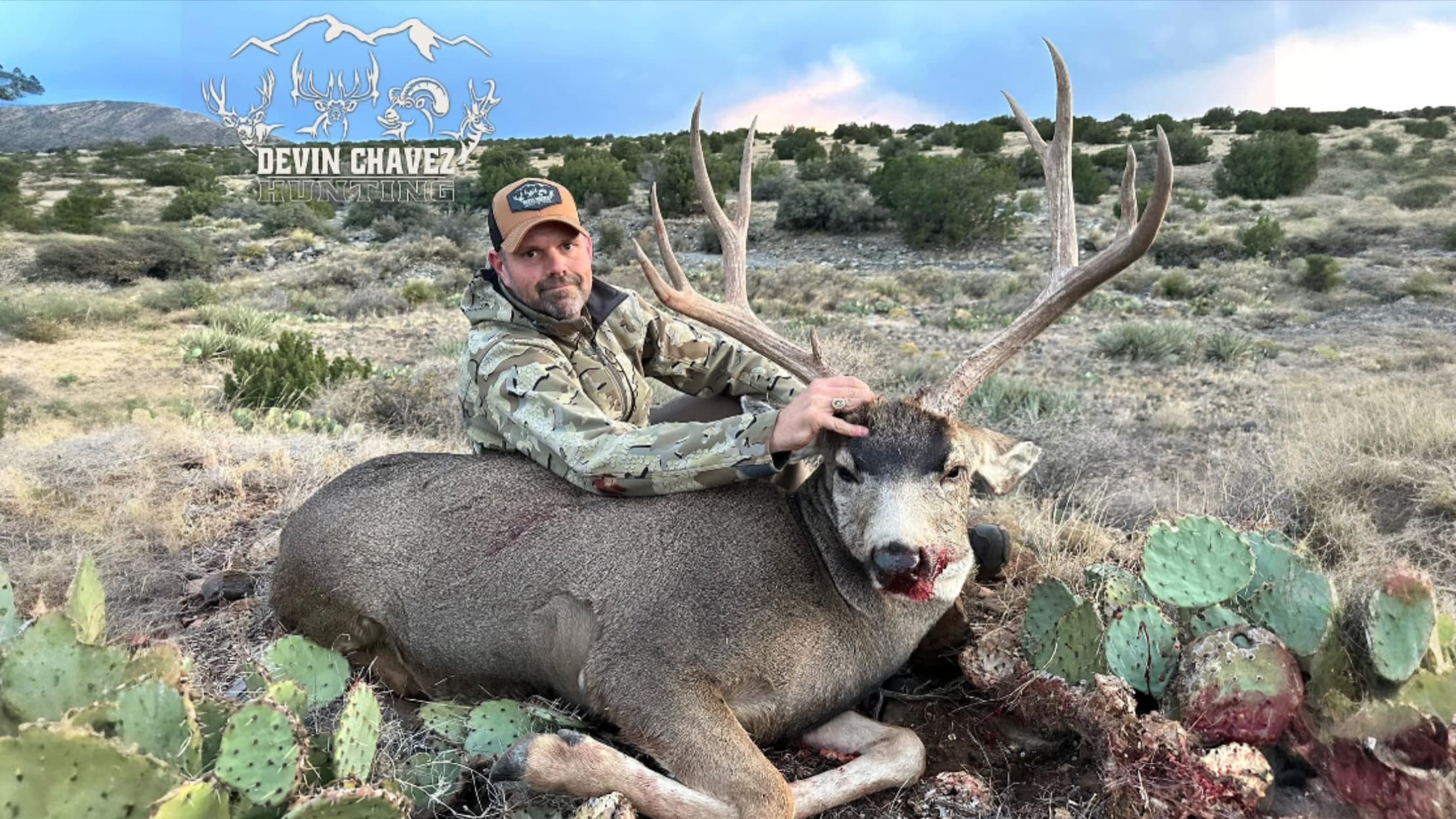 Man in camo holds deer with large antlers in field. Deer has blood on face and man is wearing a cap. Logo for "Devin Chave...