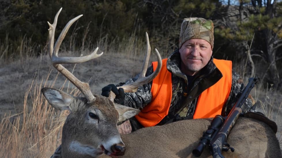 Man in hunting gear poses with a large buck deer, rifle nearby. Man wears camo, orange vest, and a camo hat. The deer has ...