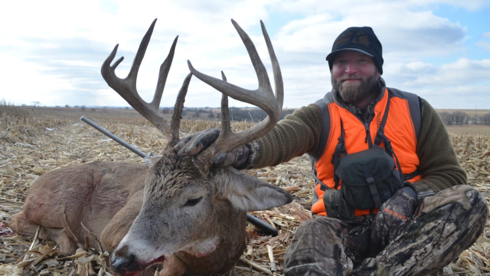 Man in hunting gear sits beside a large buck with antlers. The deer lies in a harvested corn field.