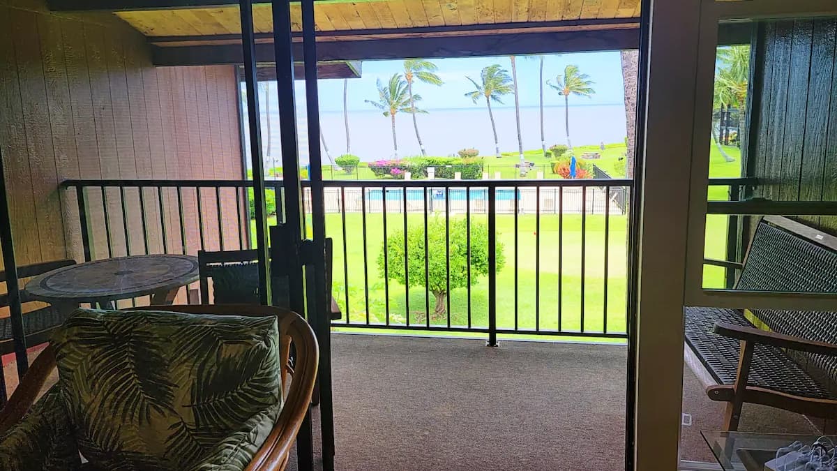 View from a balcony overlooking a lawn, a swimming pool, and the ocean with palm trees. Wicker furniture in foreground.