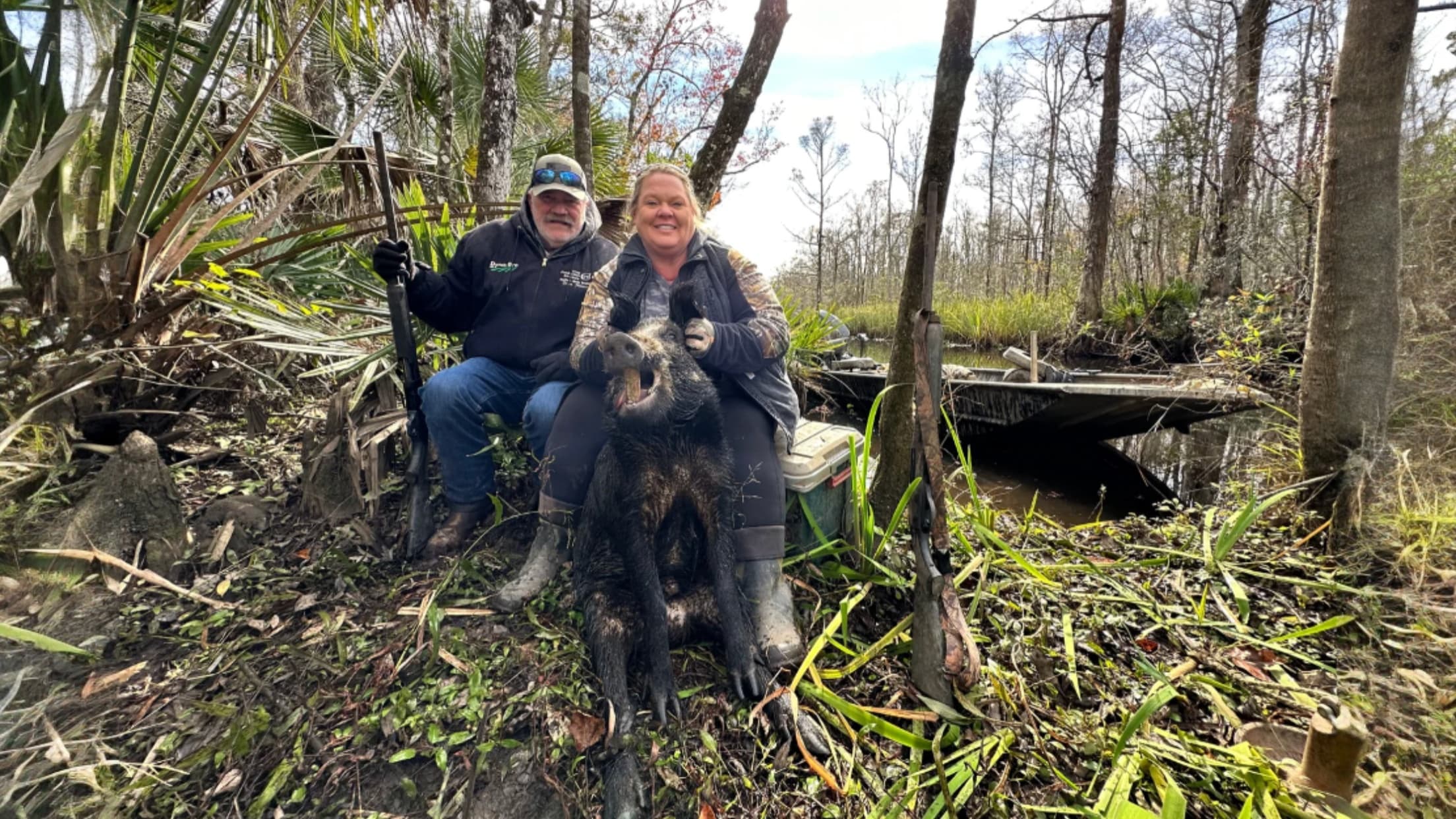 Two people posing with a large, dead wild hog. Guns and boat visible in the swampy forest environment. They are sitting in...
