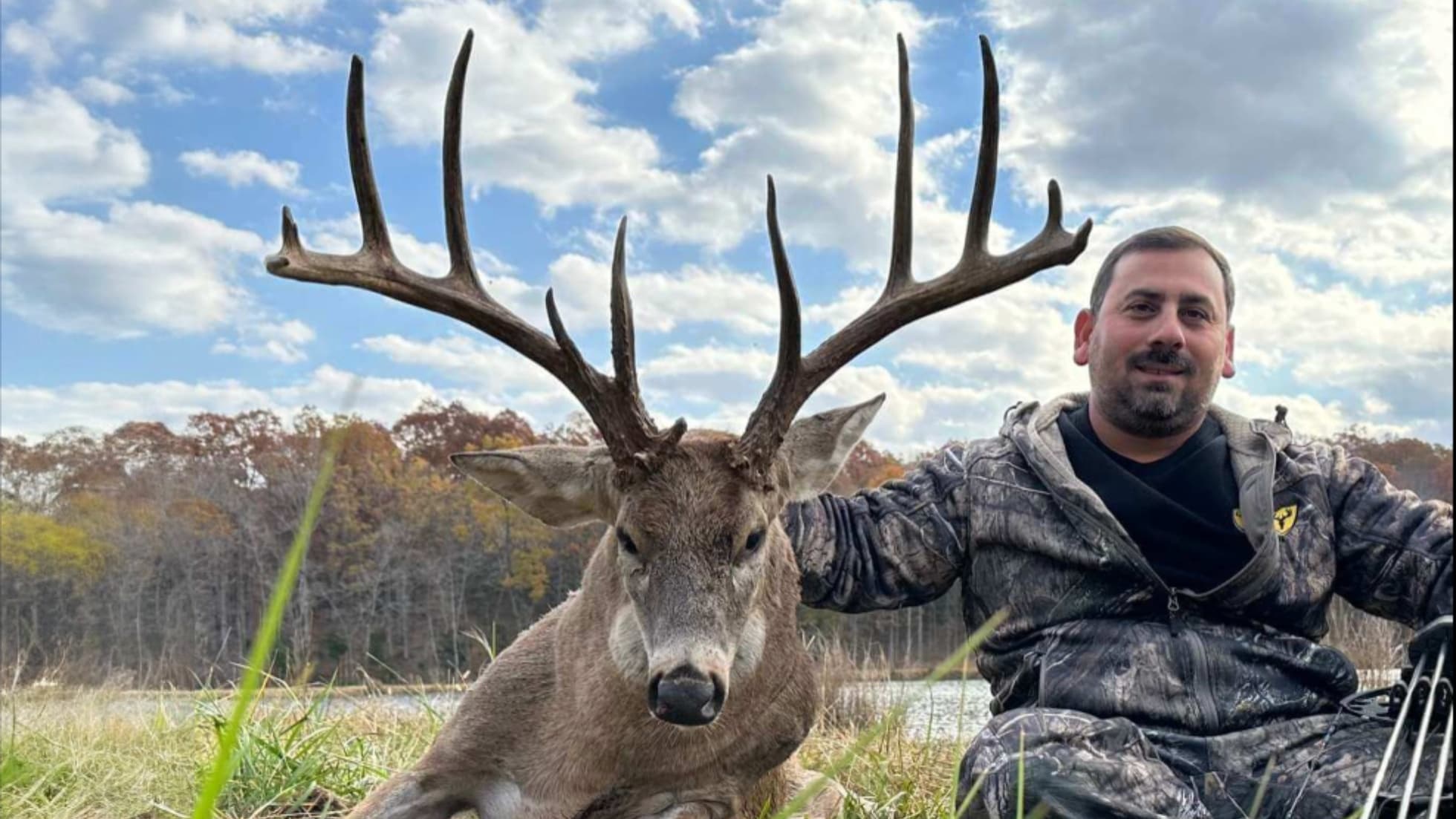 Man poses with a large buck. Deer has massive antlers. Man is wearing camo and is next to the deer outdoors. Blue sky with...