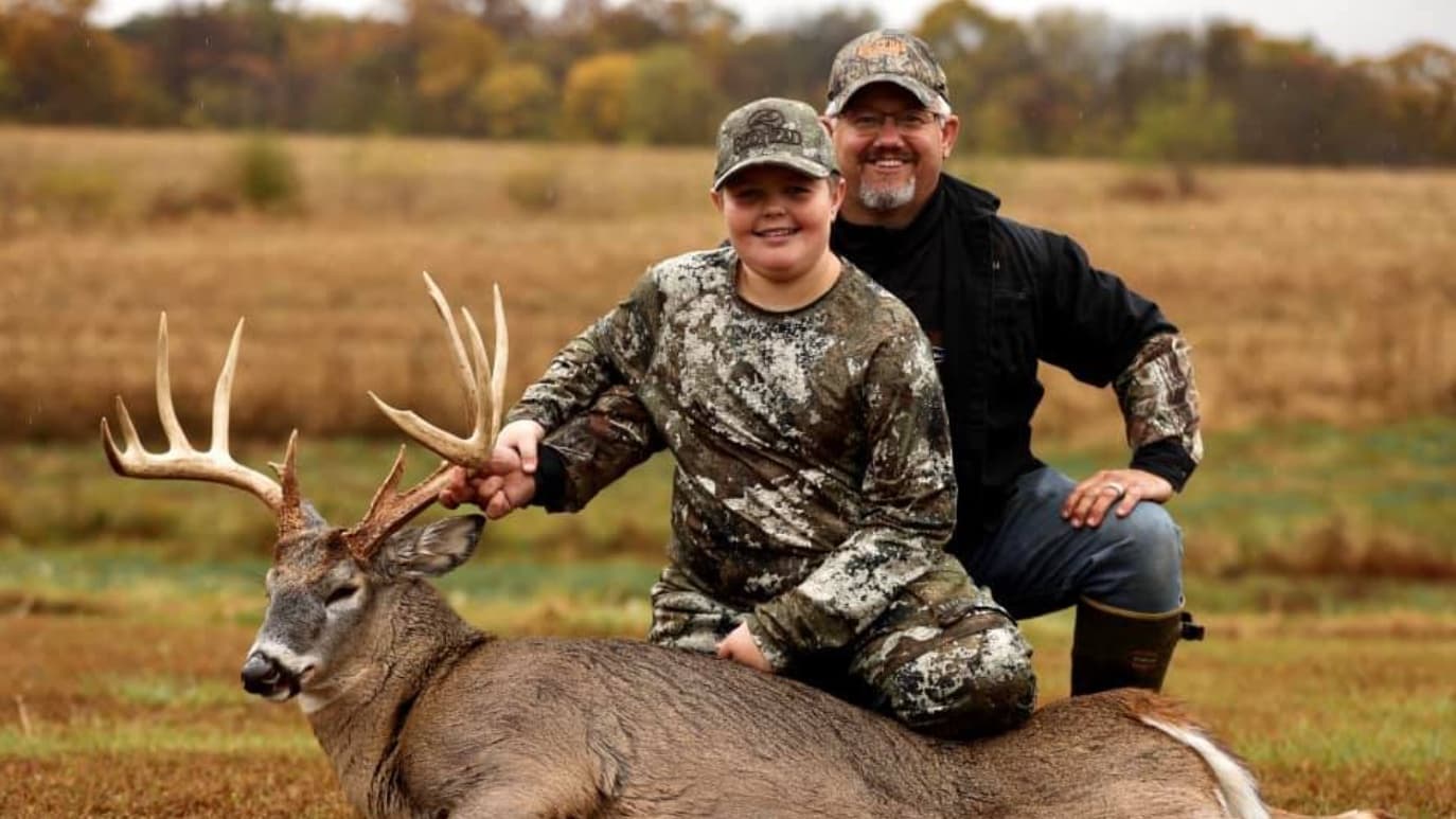 A boy in camo sits on a deer, with a man kneeling behind him. Both are smiling.  Deer antlers are prominent. Background is...
