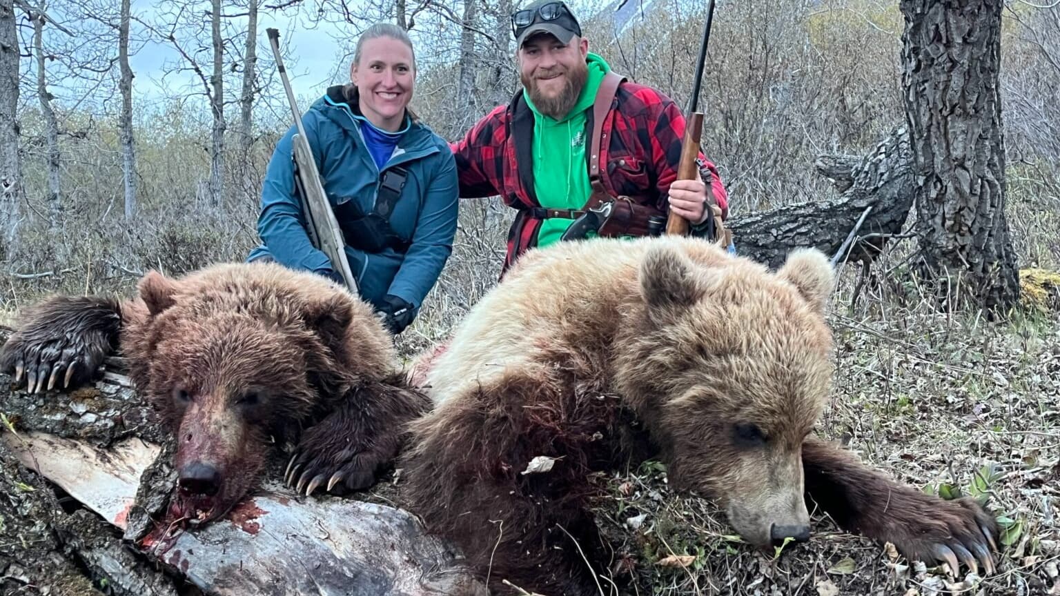 Two smiling people pose with two dead brown bears. One holds a rifle, both near a forest. The bears are lying down.