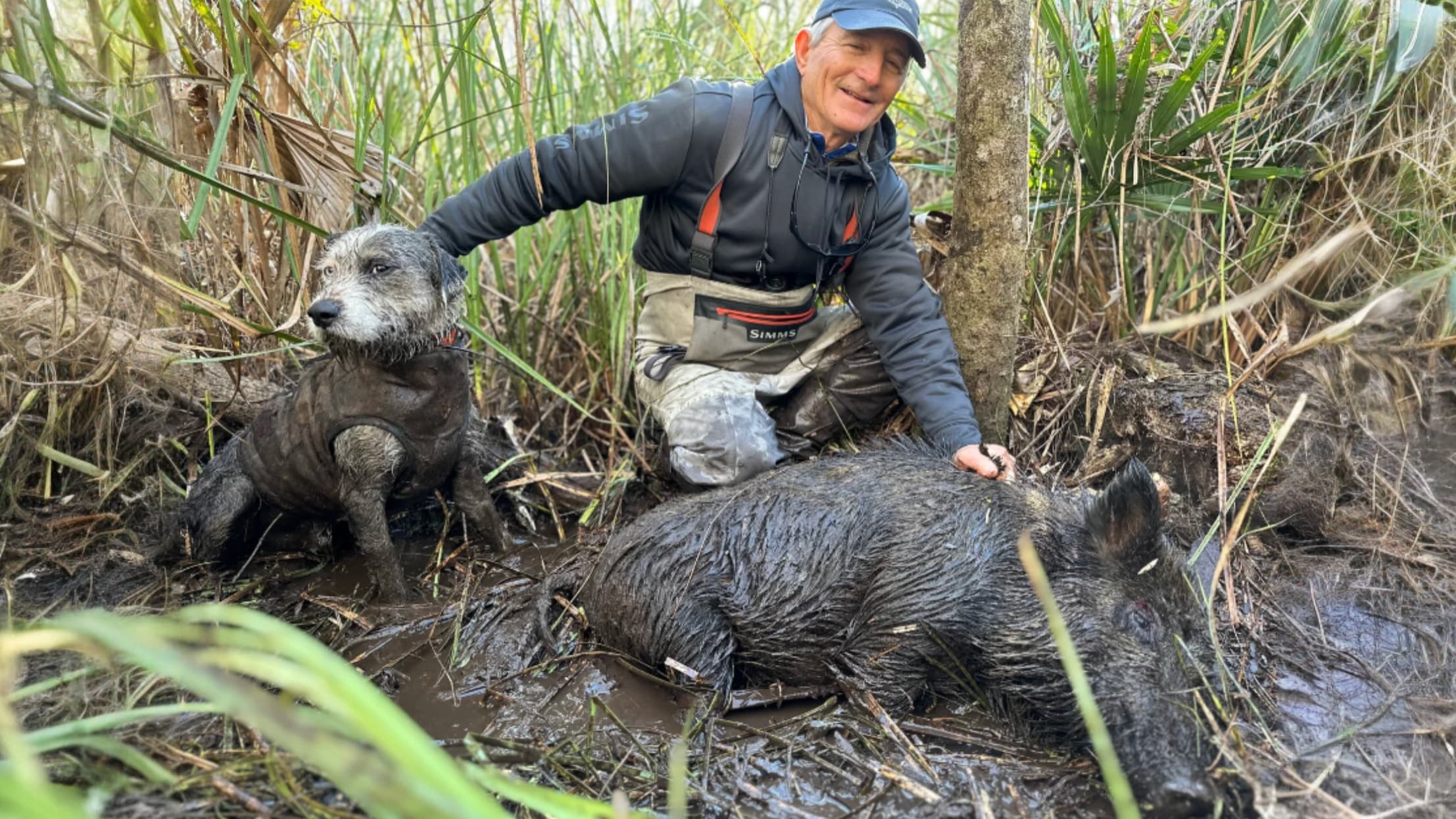 Man in waders and a small dog sit beside a wild boar in muddy water. Dog and boar appear covered in mud. Smiling man touch...
