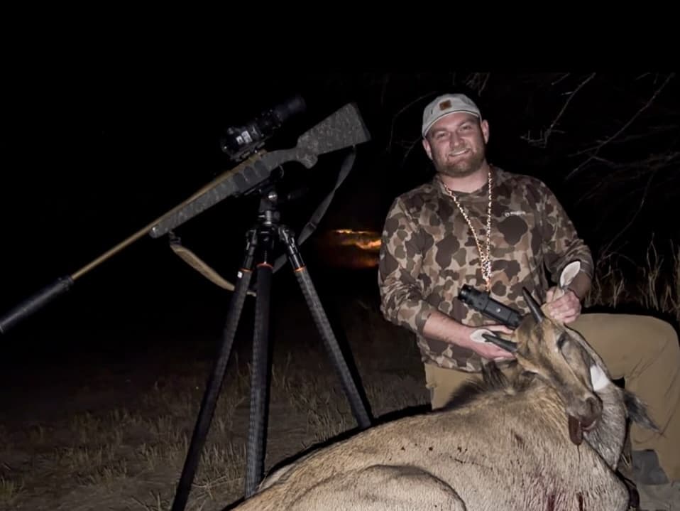 Man in camouflage holds a dead antelope, with a rifle on a tripod in the dark, near what appears to be a sunset glow.