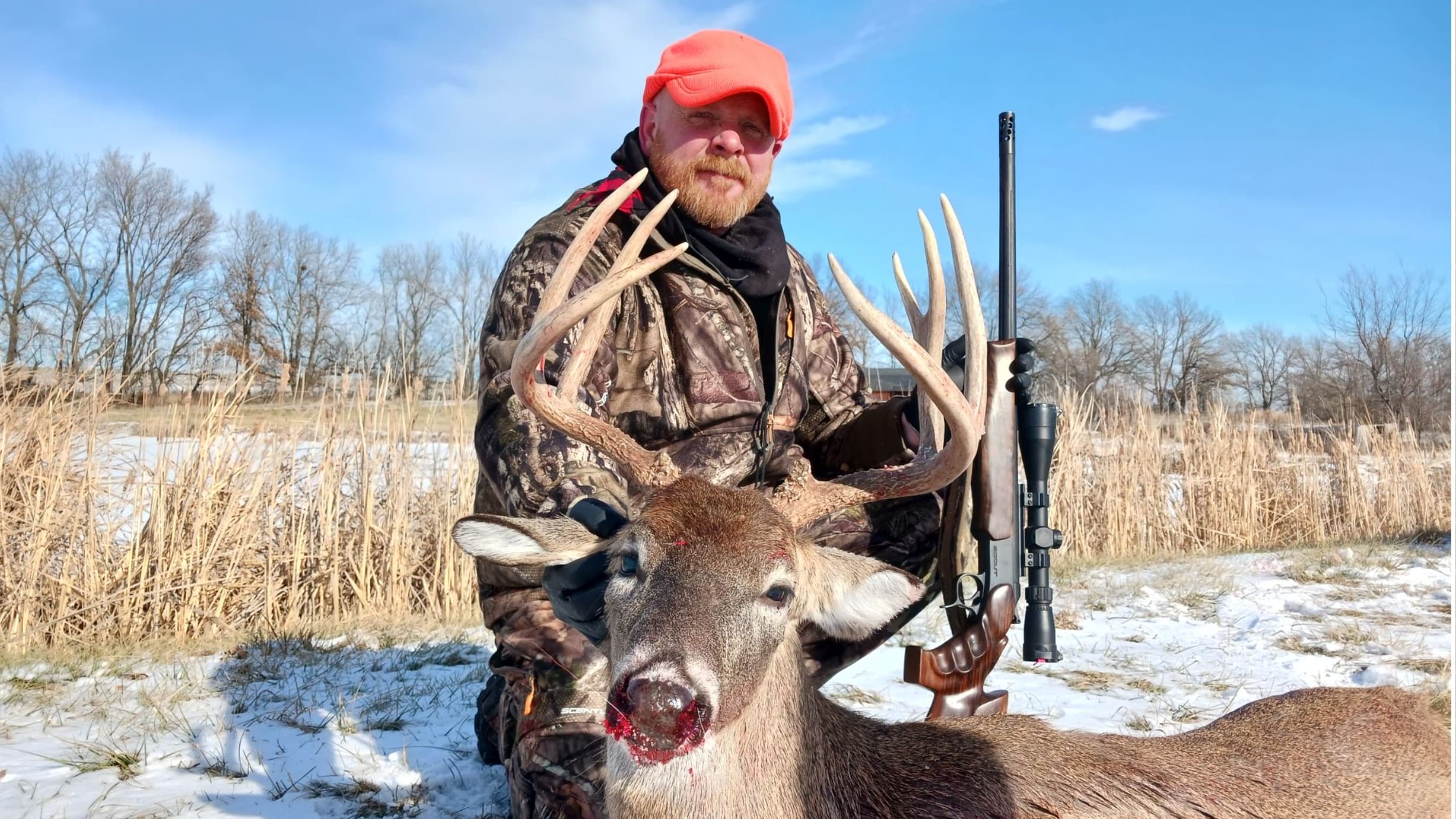 Hunter in camouflage and orange hat holds a large buck's antlers. Rifle stands beside him. Snow and dry grass surround them.