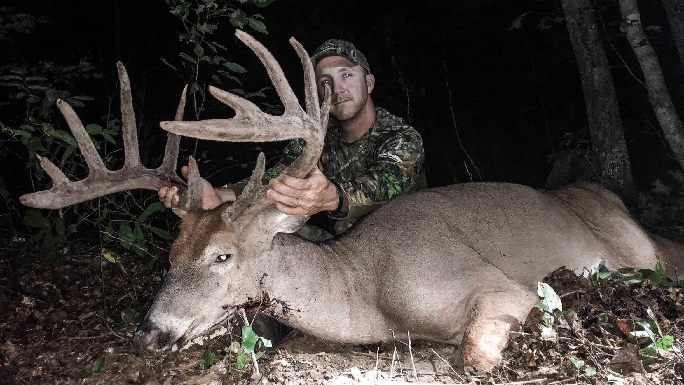 Hunter in camouflage holds large antler of a buck lying on forest floor. Deer has a wound. Dark background, forest setting.