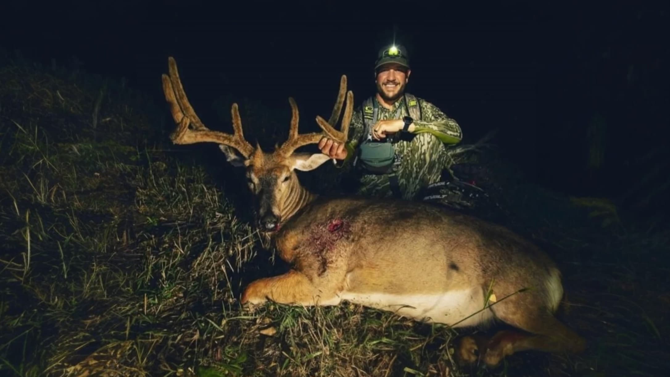 Hunter smiles next to a large buck lying on the grass. The buck has large antlers, a wound on its side, and is in low ligh...