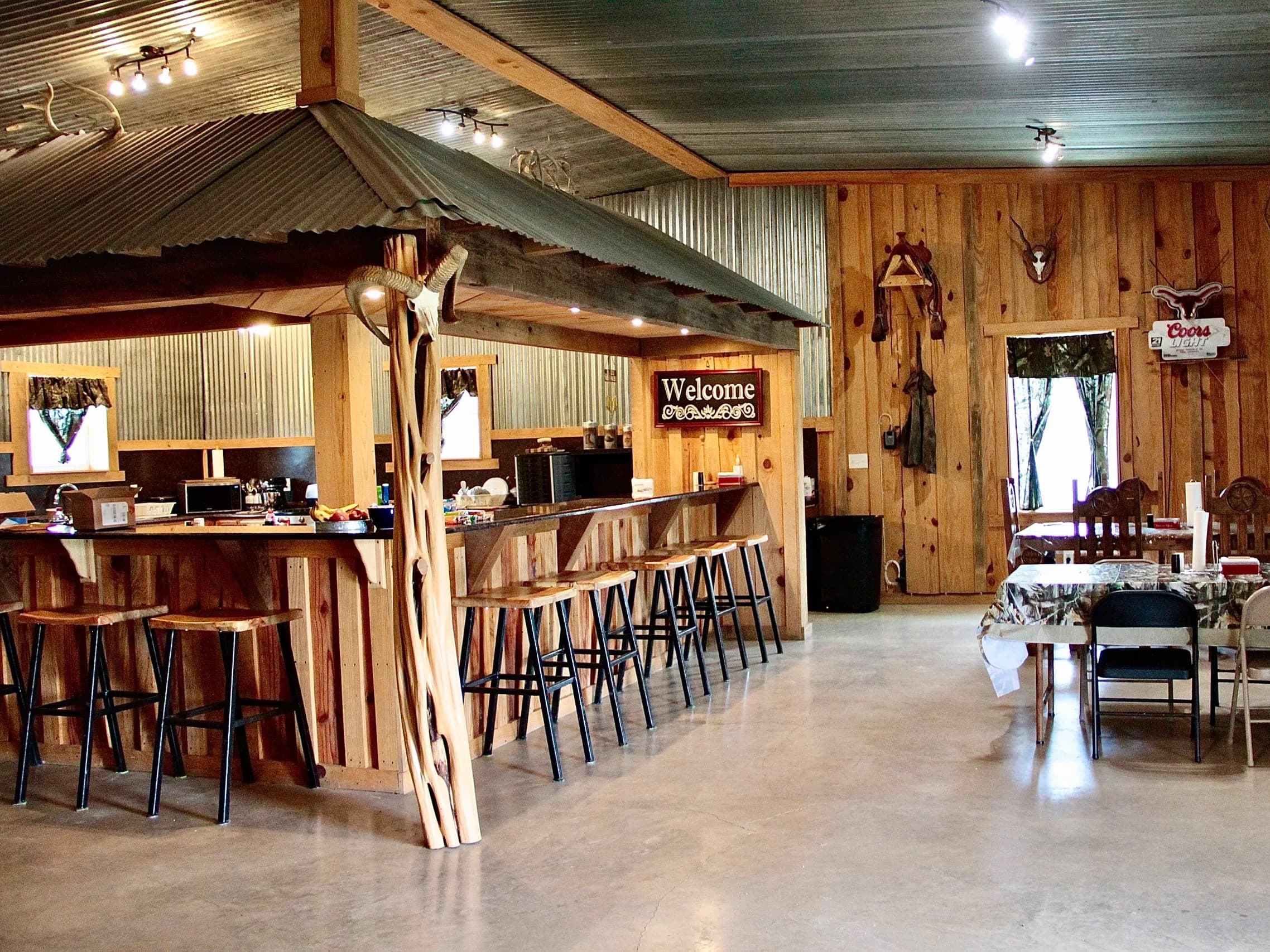 Rustic bar with a corrugated metal roof, wooden stools, and "Welcome" sign. Deer antlers and Coors Light sign adorn the wo...