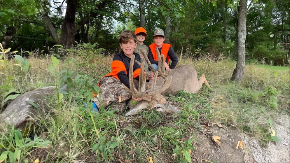 Three boys in hunter's orange vests pose with a large buck. The deer lies in grass, displaying large antlers. Trees in bac...