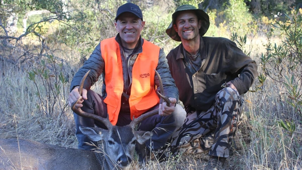Two smiling men kneeling beside a harvested buck. One wears an orange hunting vest, holding antlers. The other in camoufla...