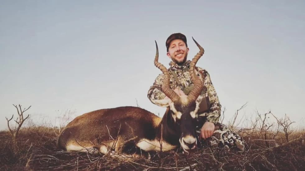 Man in camo smiles next to a blackbuck on the ground. The blackbuck has large spiraling horns. Grassy field under blue sky.