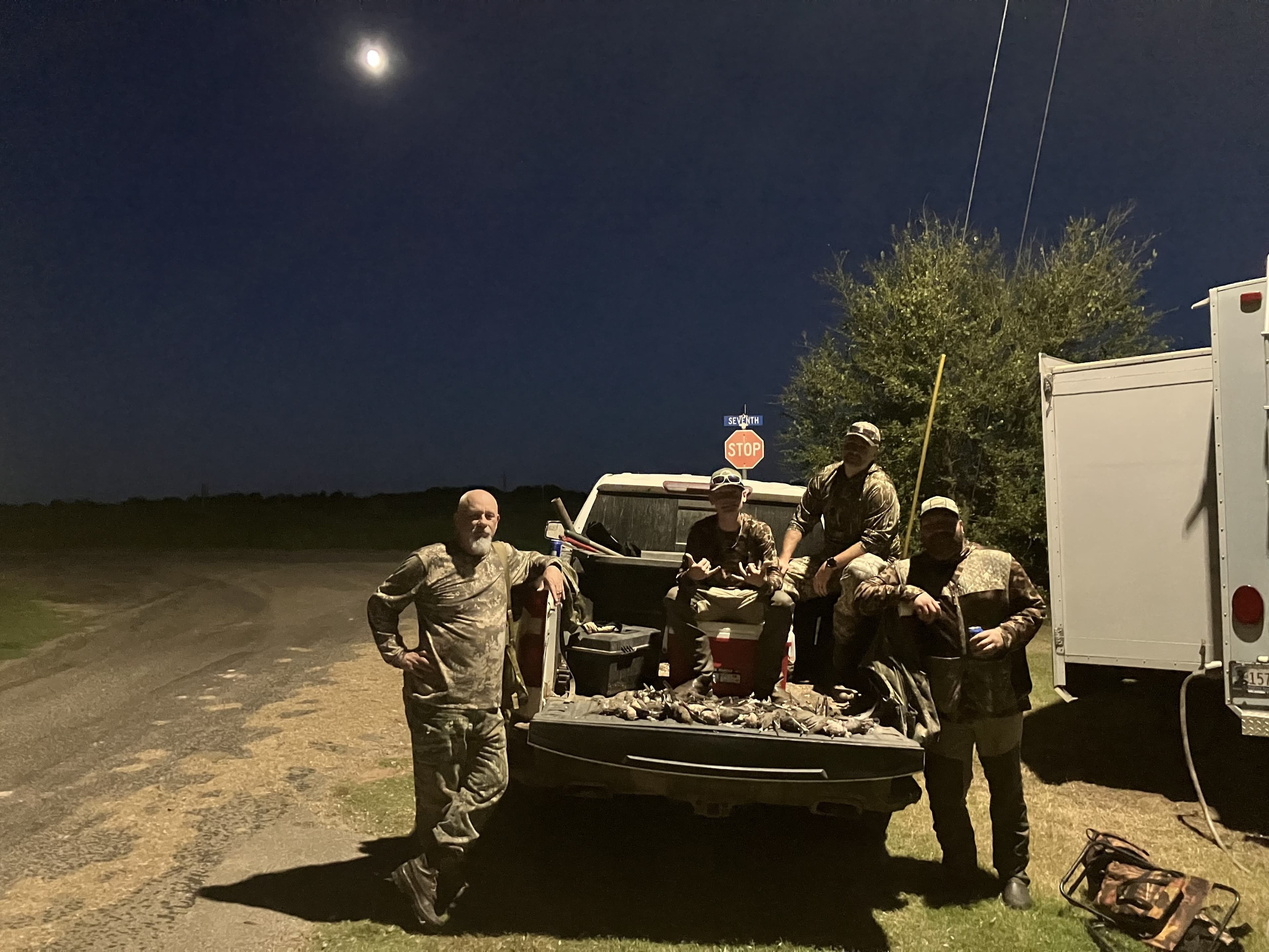 Group of hunters gathered at night, posing by a pickup truck with a large harvest. Moon and road sign visible in backgroun...