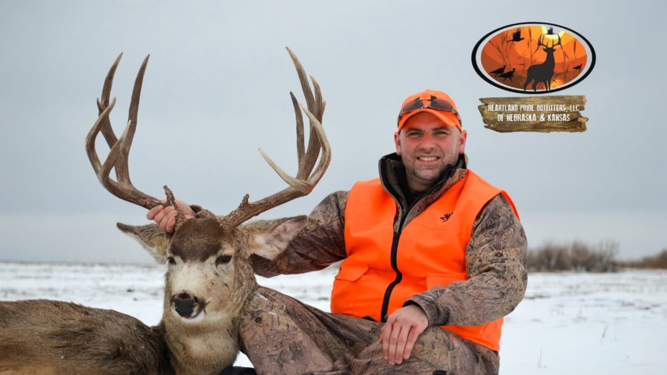 Man in orange vest and hat with large-antlered deer on snow. Heartland Pride Outfitters logo in upper right.
