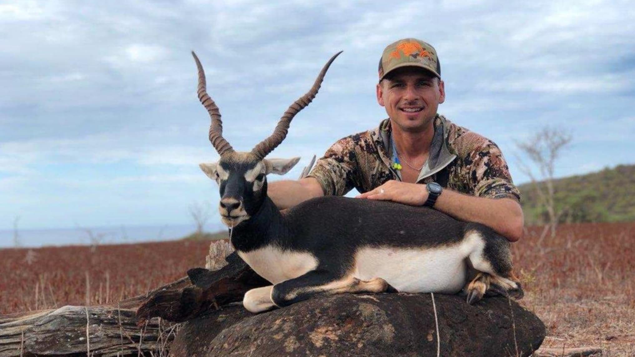 Man poses with blackbuck antelope; man pats antelope lying on a rock. Antelope has large spiraled horns, white underbelly....