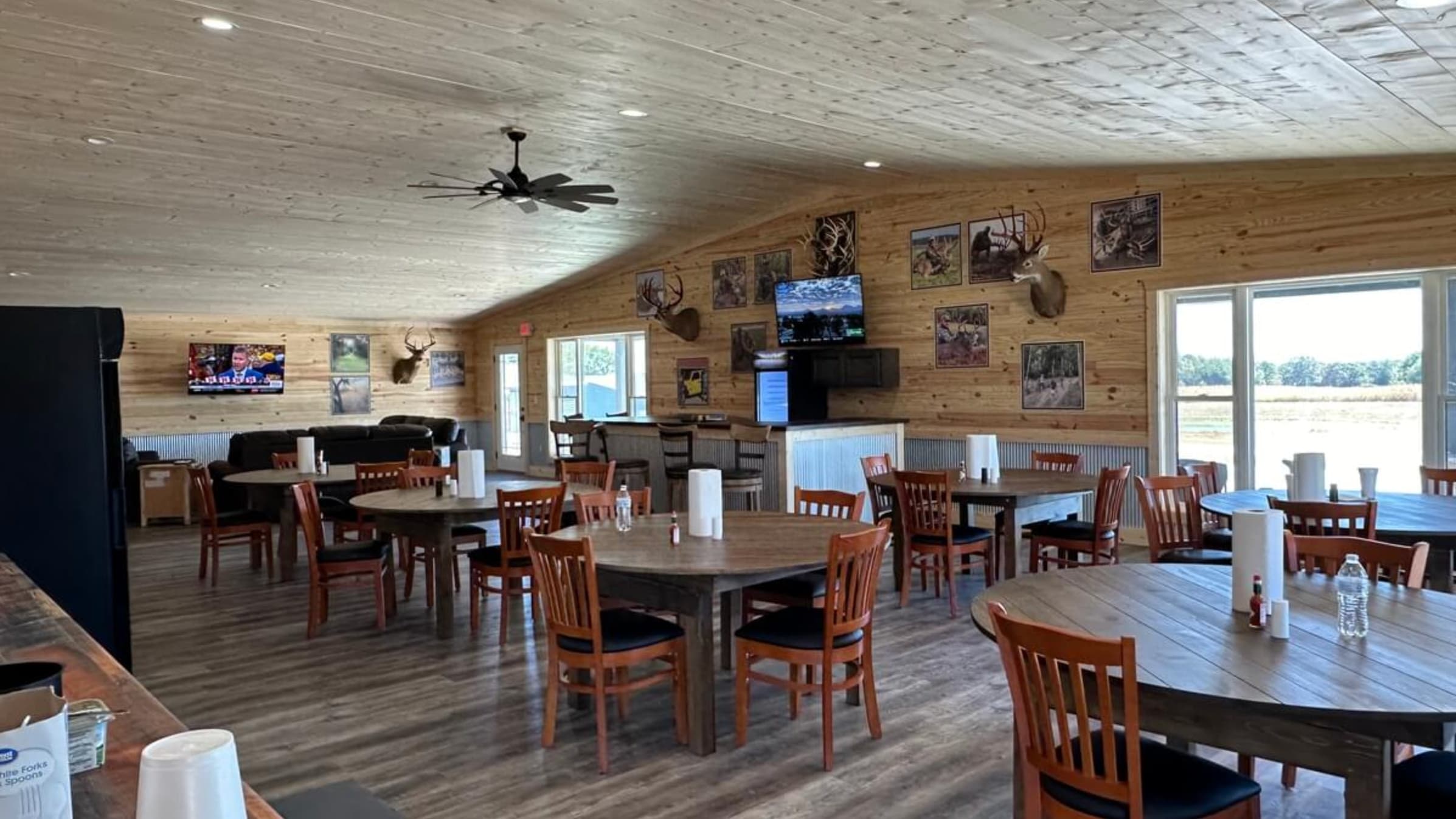 Interior of a hunting lodge with wood paneled walls, round tables, chairs, bar, and deer mounts. Large windows offer an ou...