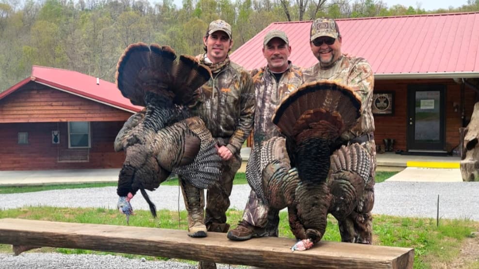 Three men in camouflage holding two large turkeys. The men stand in front of a wooden building with a red roof.