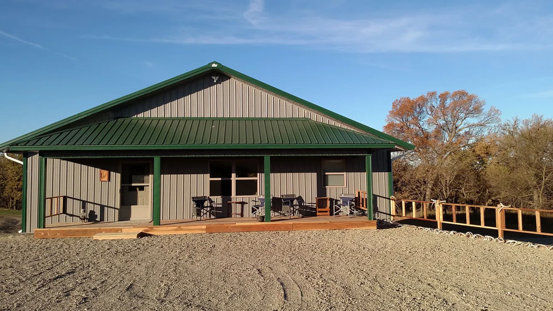 A modern, gray metal-clad building with a green roof and porch. Several chairs and a grill are set up. Gravel yard, clear ...