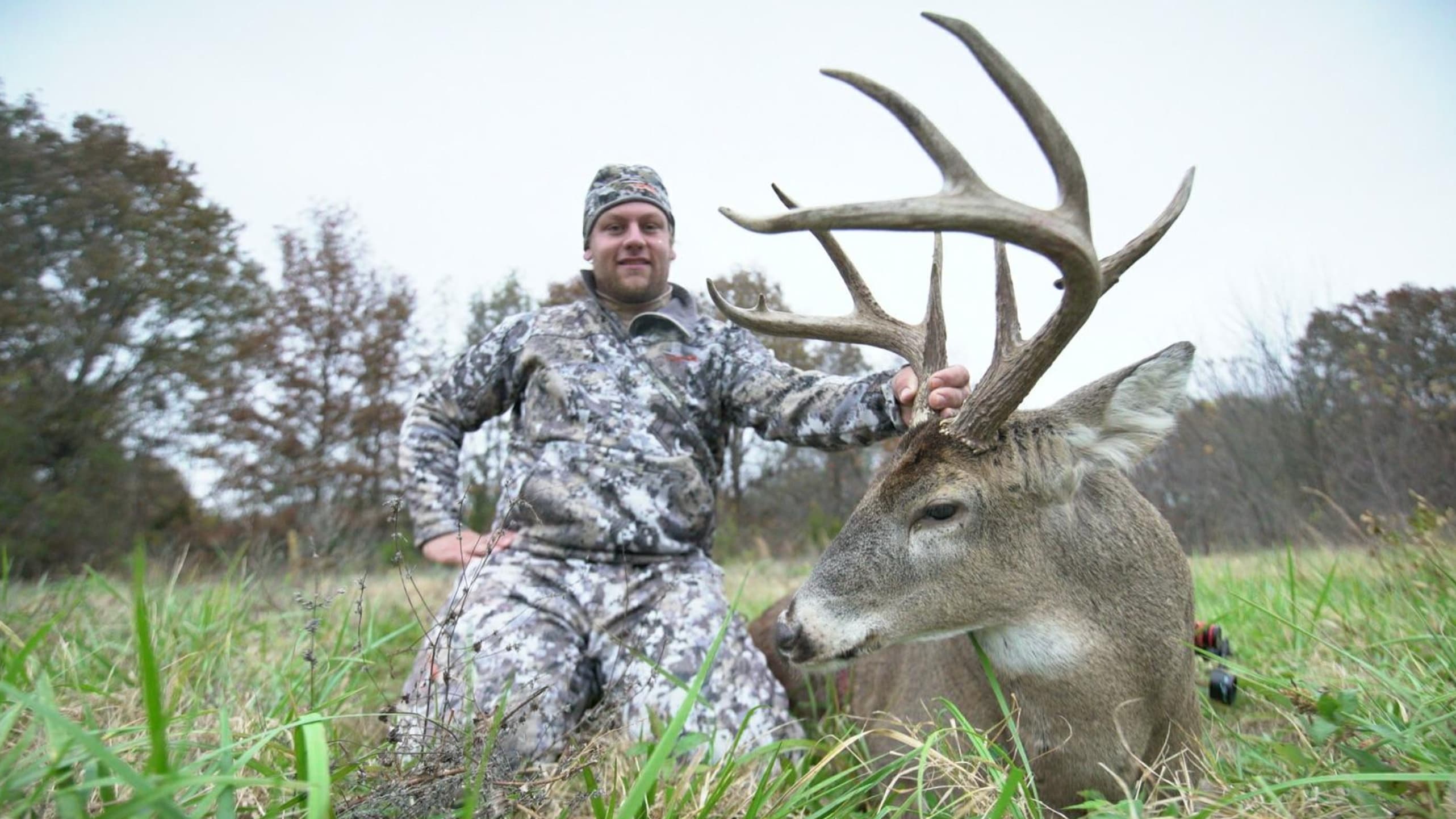 Hunter in camouflage kneels next to a large buck with impressive antlers. Grassy foreground, trees in the blurred background.