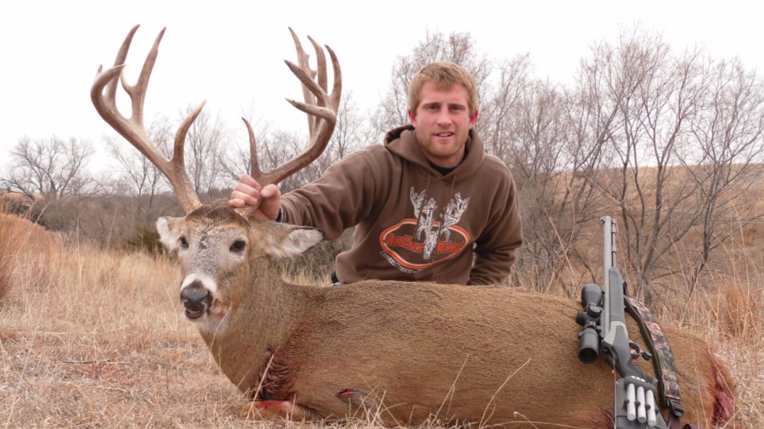 A man in a brown hoodie stands next to a large buck with impressive antlers, lying on the ground. A rifle rests nearby.