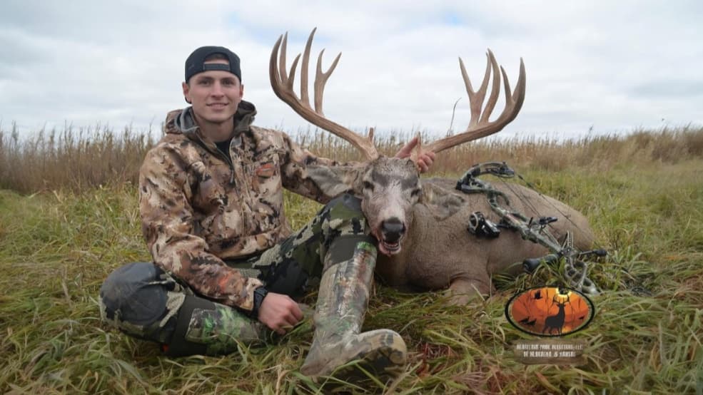 Hunter poses with large buck with impressive antlers. He sits beside the deer with a bow and arrow lying next to the anima...