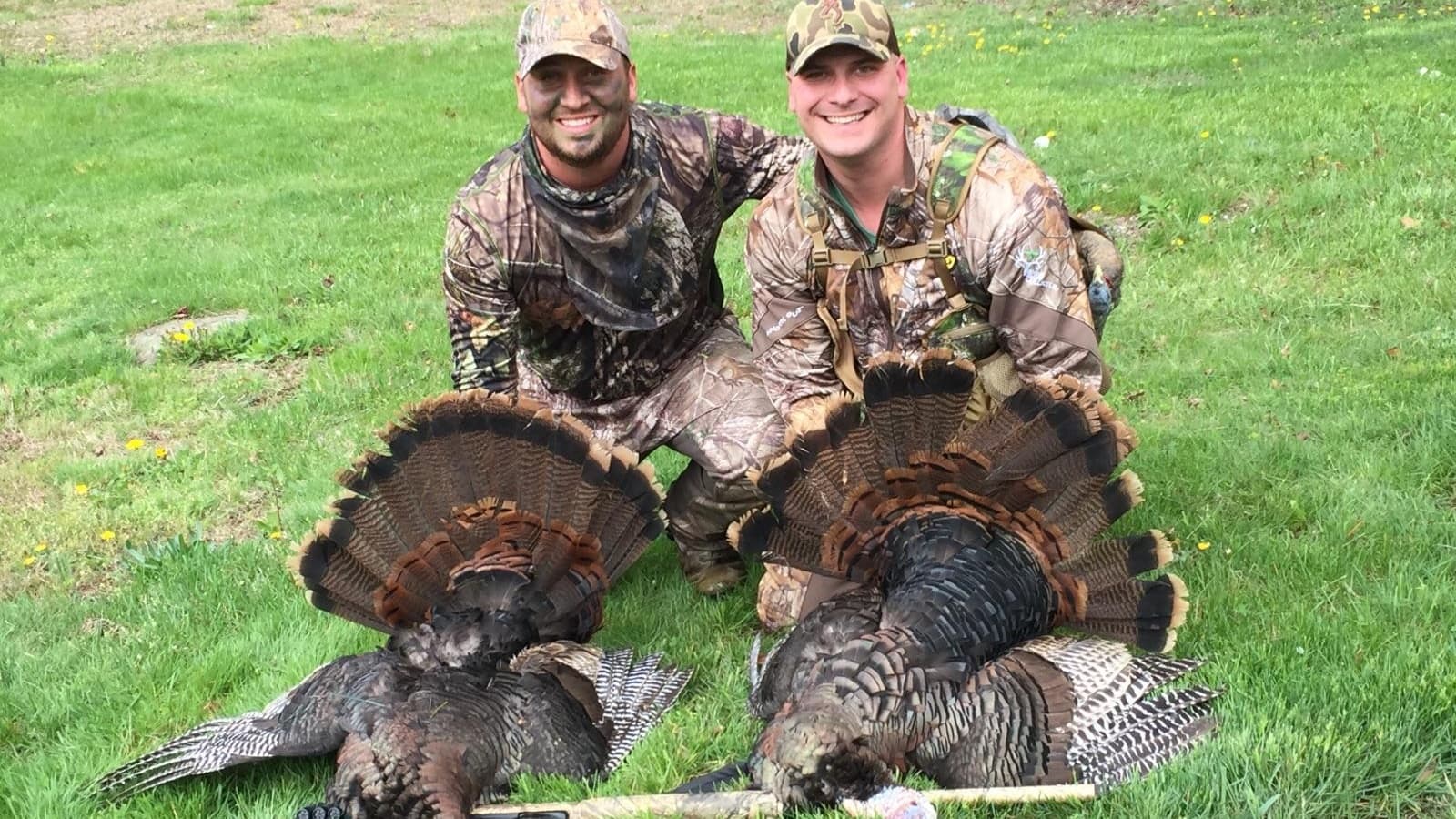Two men in camouflage pose next to two dead turkeys with fanned-out tail feathers on a grassy field. Both men are smiling.