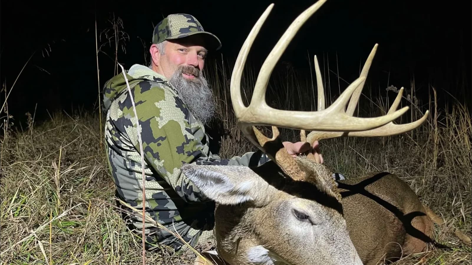 Man in camo hat and jacket kneels beside a large buck, holding its impressive antlers. Background is dark with tall grass.