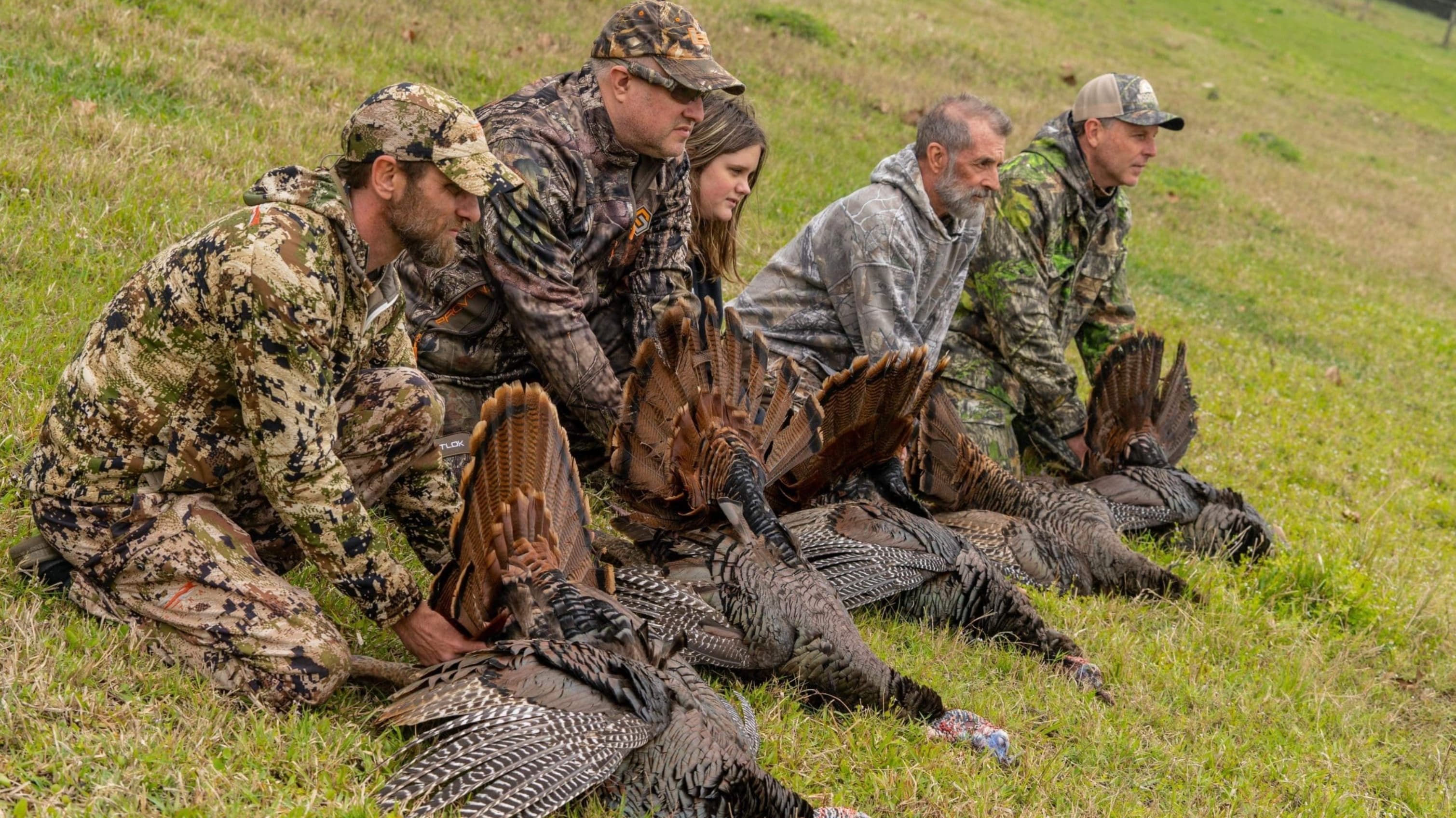 A group of hunters in camouflage kneel beside harvested turkeys with fanned tail feathers, displayed on a grassy field.