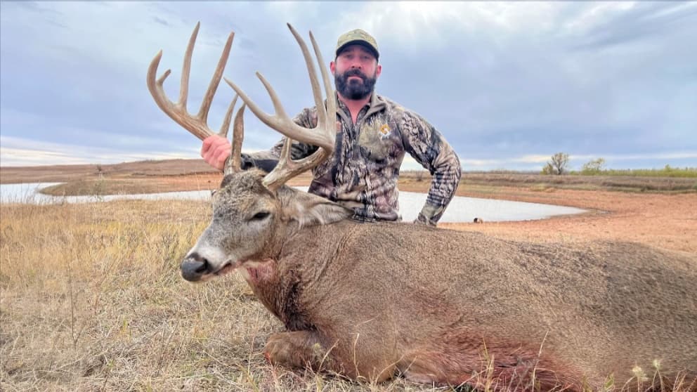 Man in camouflage stands with a large-antlered buck he has hunted. The deer lies in tall grass, a small body of water in t...