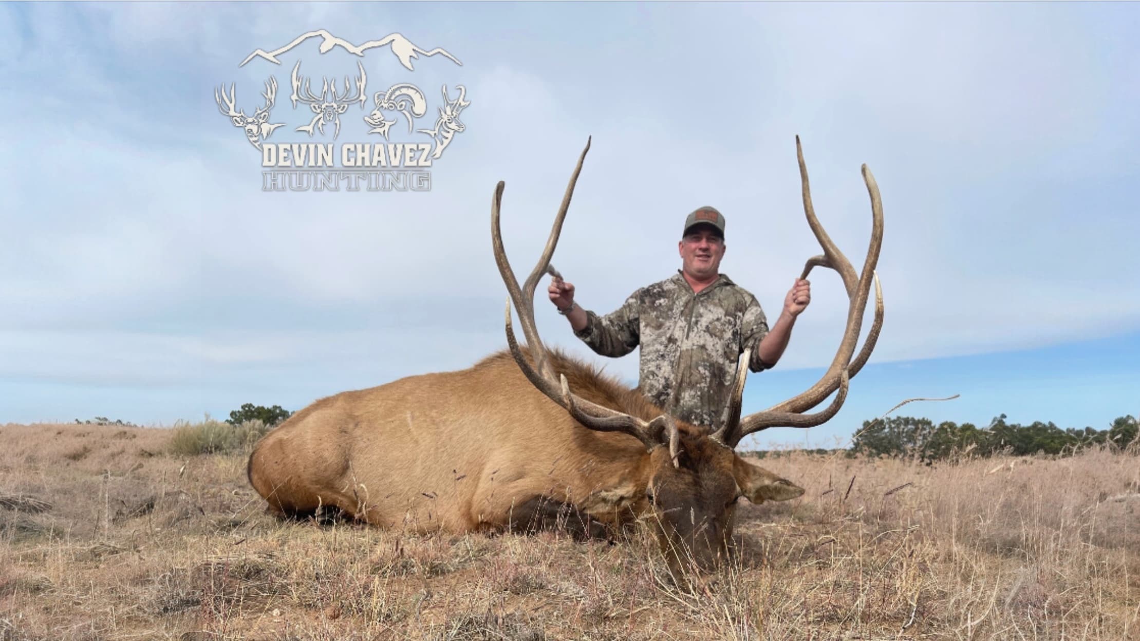 Man holding large elk antlers next to a harvested elk. Camo clothing. Field and blue sky background. "Devin Chavez Hunting...