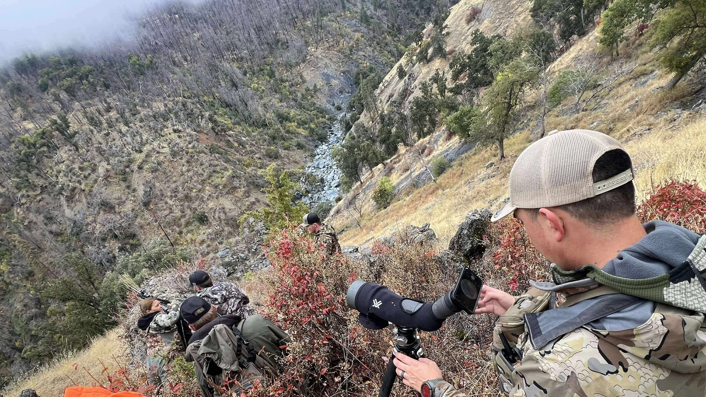 Hunters on a steep, rugged mountainside. One man looks through a spotting scope. Others are camouflaged, observing the val...