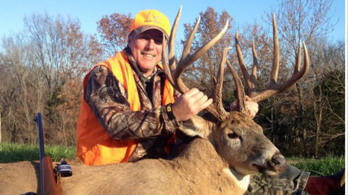 Hunter in orange vest proudly holds large rack deer. Rifle visible.
