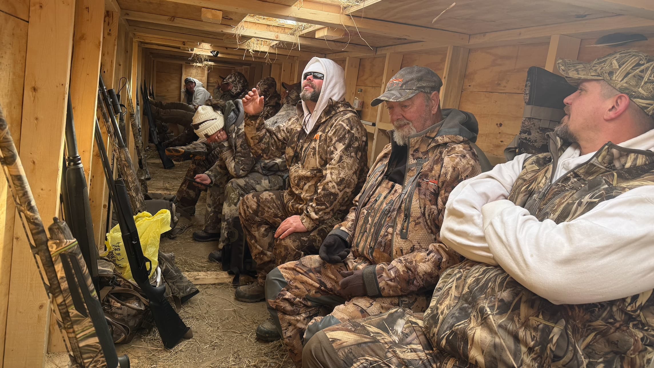 Group of hunters in camouflage clothing inside a wooden hunting blind. Shotguns line the walls. Men sit and look out.