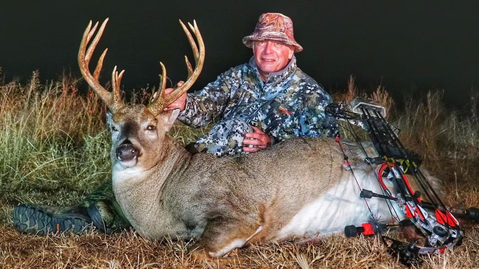 Man in camo hat and jacket poses with a large buck. The buck lays on the ground, a bow rests beside it.