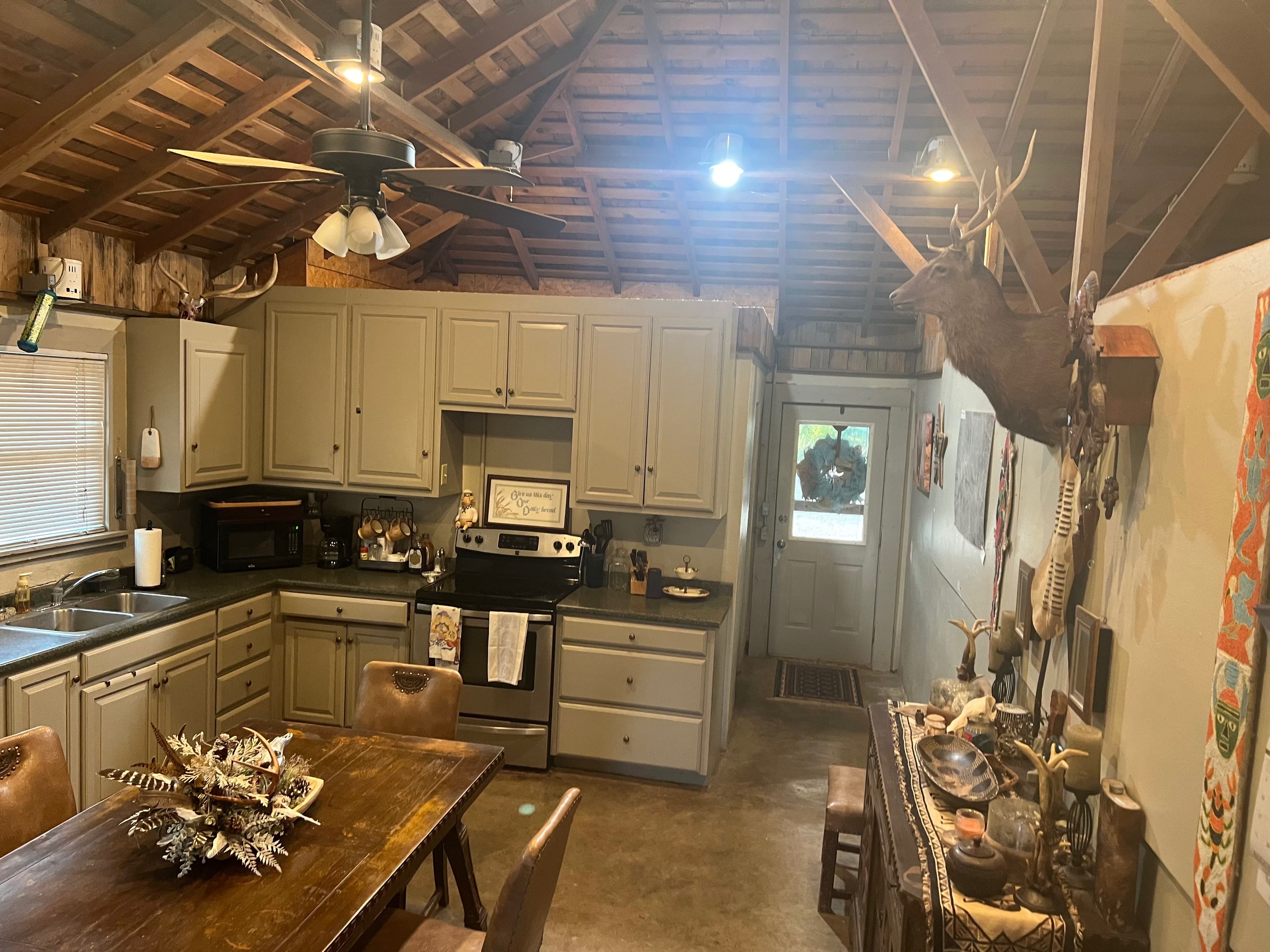 Rustic kitchen with wood ceiling, cabinets, and a table. Antlers and taxidermy deer head adorn walls. Door leads outside.