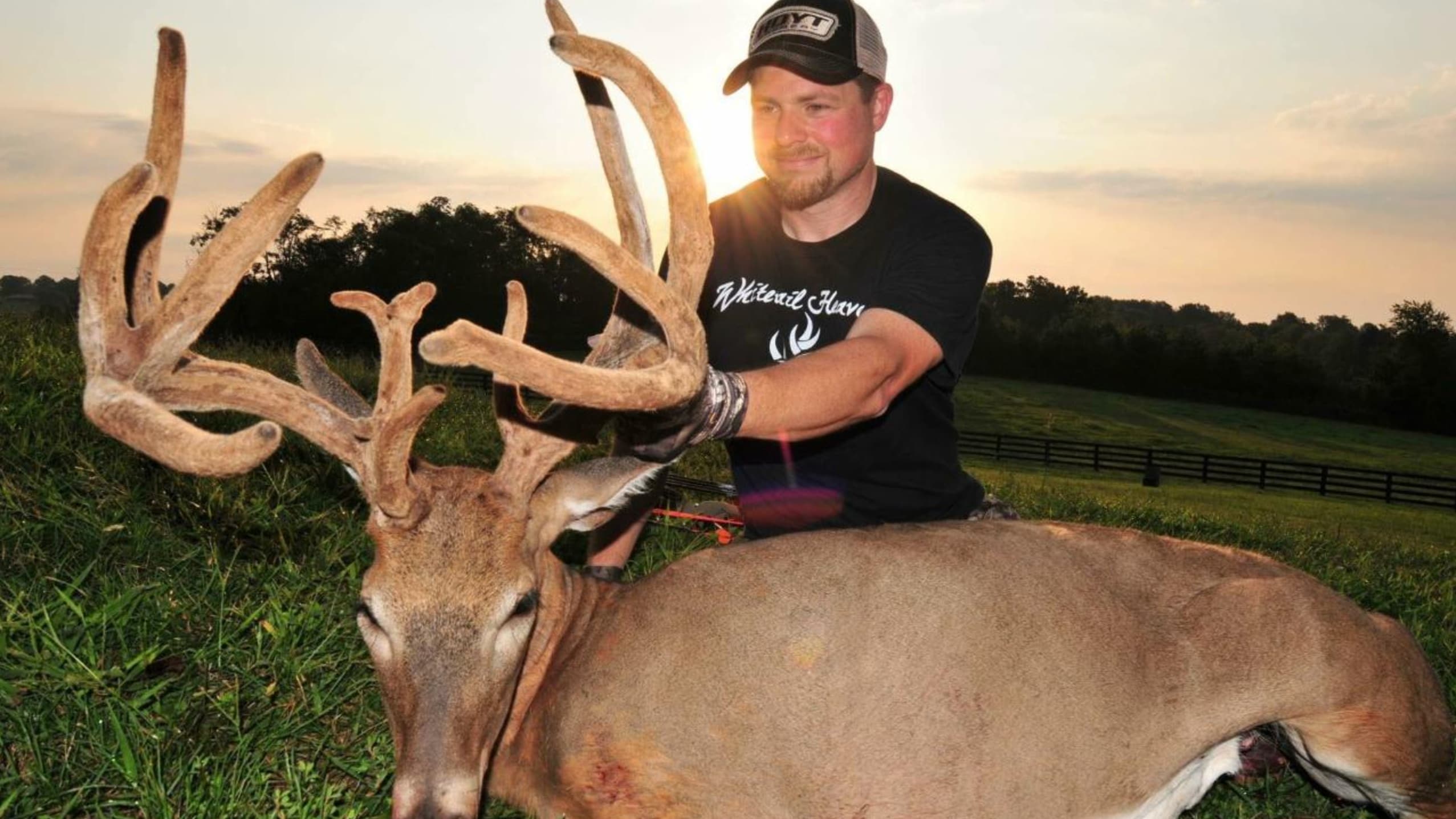 Man kneeling beside a large dead deer with impressive antlers. Deer lies in grass. Man wears black shirt, cap, gloves. Sky...