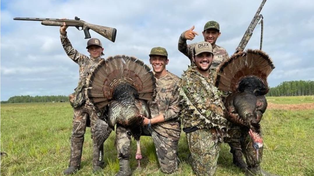 Four hunters in camouflage with shotguns pose with harvested turkeys in a field. One hunter holds a gun aloft.