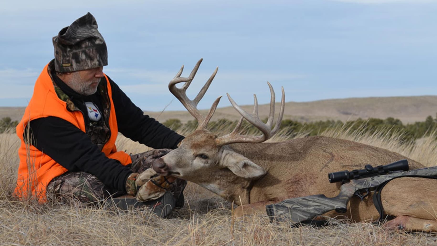 Hunter in orange vest sits next to a large buck with impressive antlers. Rifle and gear beside the deer on the ground.