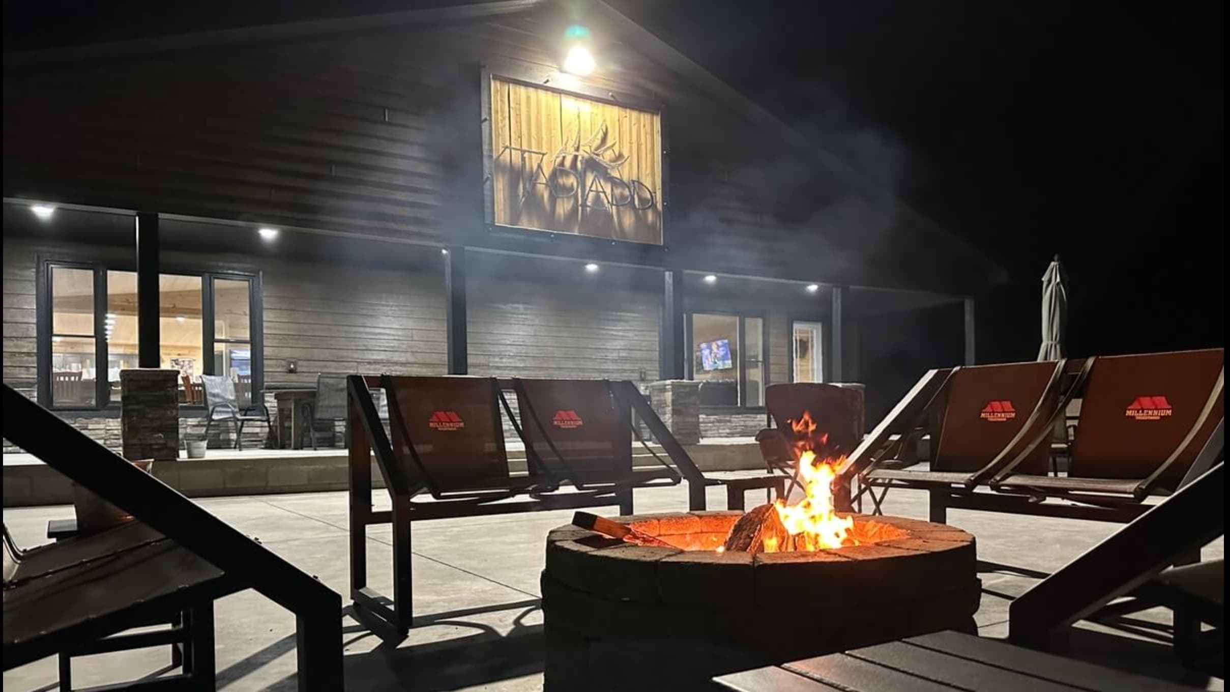 A nighttime scene with a fire pit surrounded by lounge chairs. A building with a sign and lights is in the background. Fir...