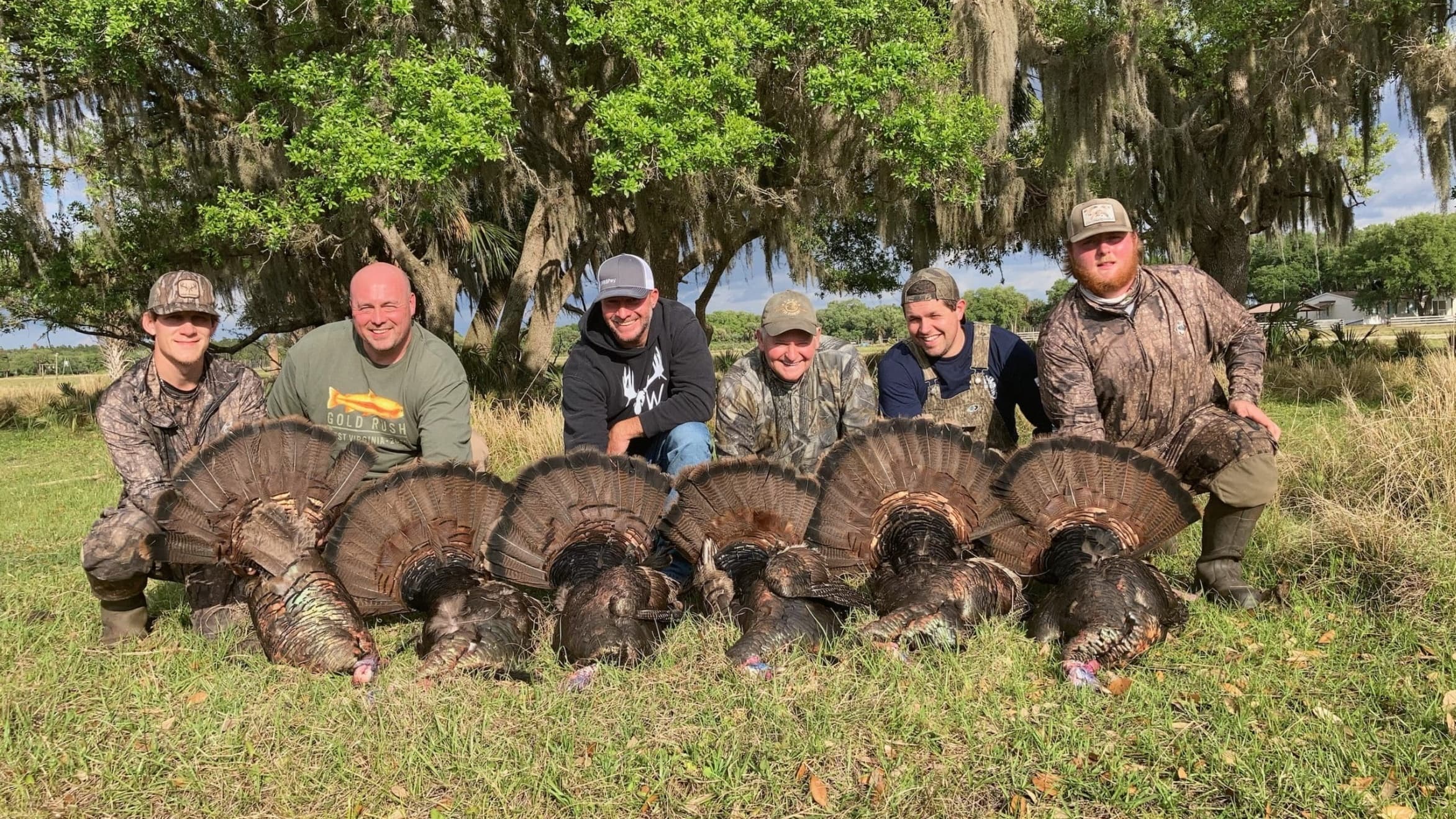 Group of six men with six harvested turkeys on grass, posing for a photo under a tree with Spanish moss.
