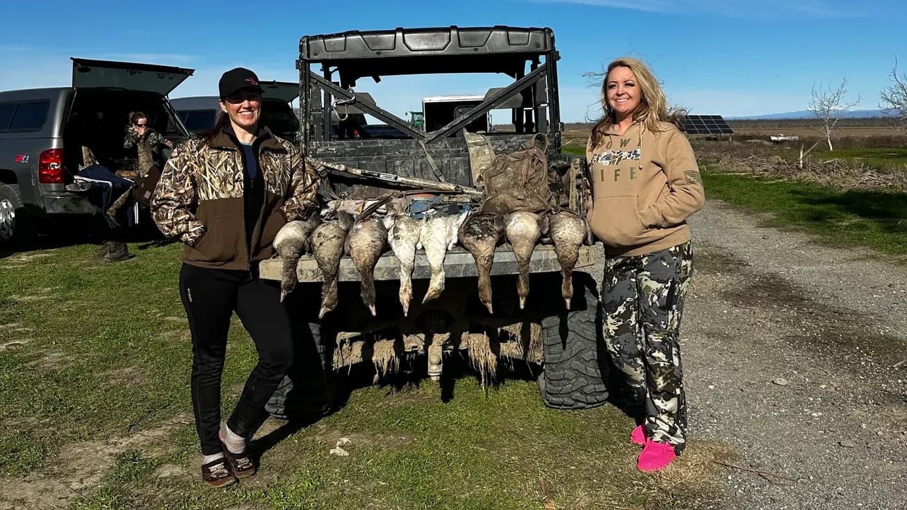 Two women stand next to an off-road vehicle with harvested ducks hanging from it. One person is wearing camo. Clear, sunny...