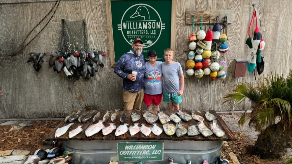 Three people pose in front of a sign and a display of caught fish. Fishing net with ducks hangs above. Colorful buoys deco...