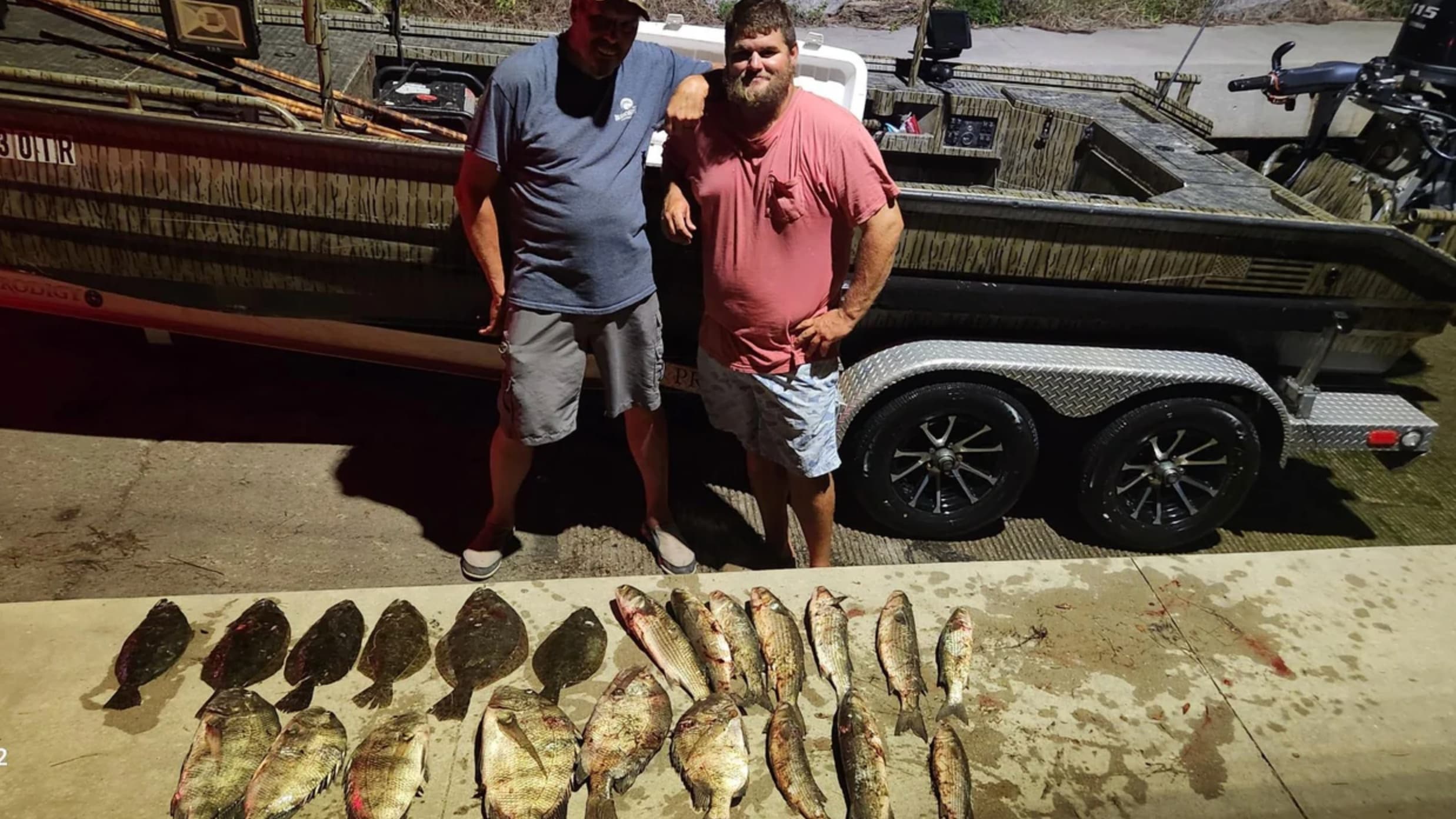 Two men pose by a camouflage boat and a concrete surface with numerous fish. Fish are laid out. The boat is on a trailer.