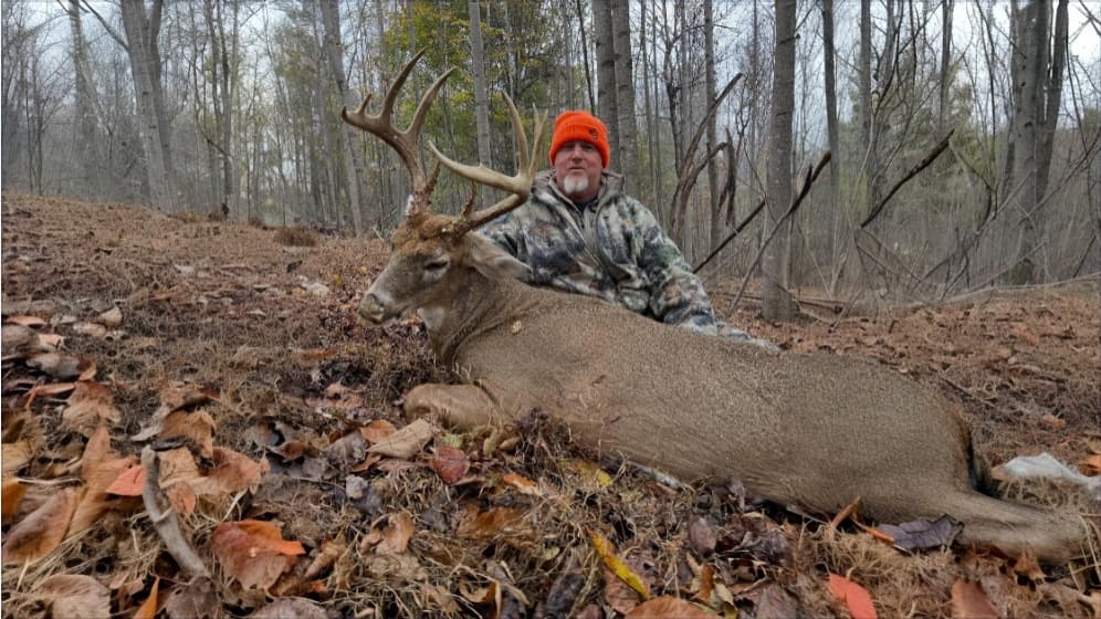 Man in camouflage and orange hat poses with a large buck with antlers lying on the forest floor, surrounded by leaves.