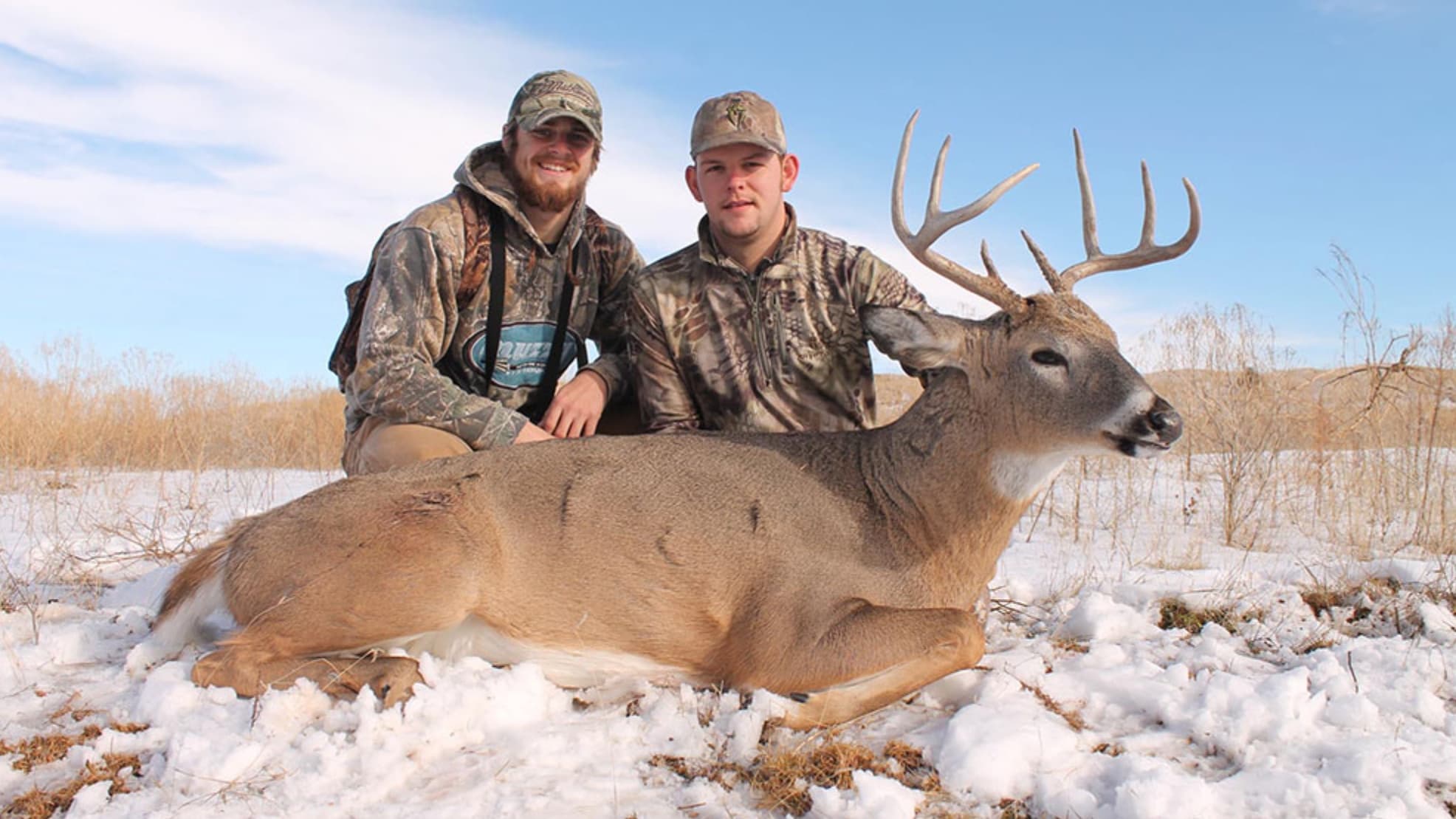 Two men in camouflage pose with a large buck deer, lying in snow. The deer has large antlers, resting in a winter landsca...