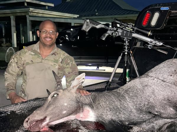 Man in camo shirt with a downed deer. Rifle on tripod in the background. Deer lying on a surface.
