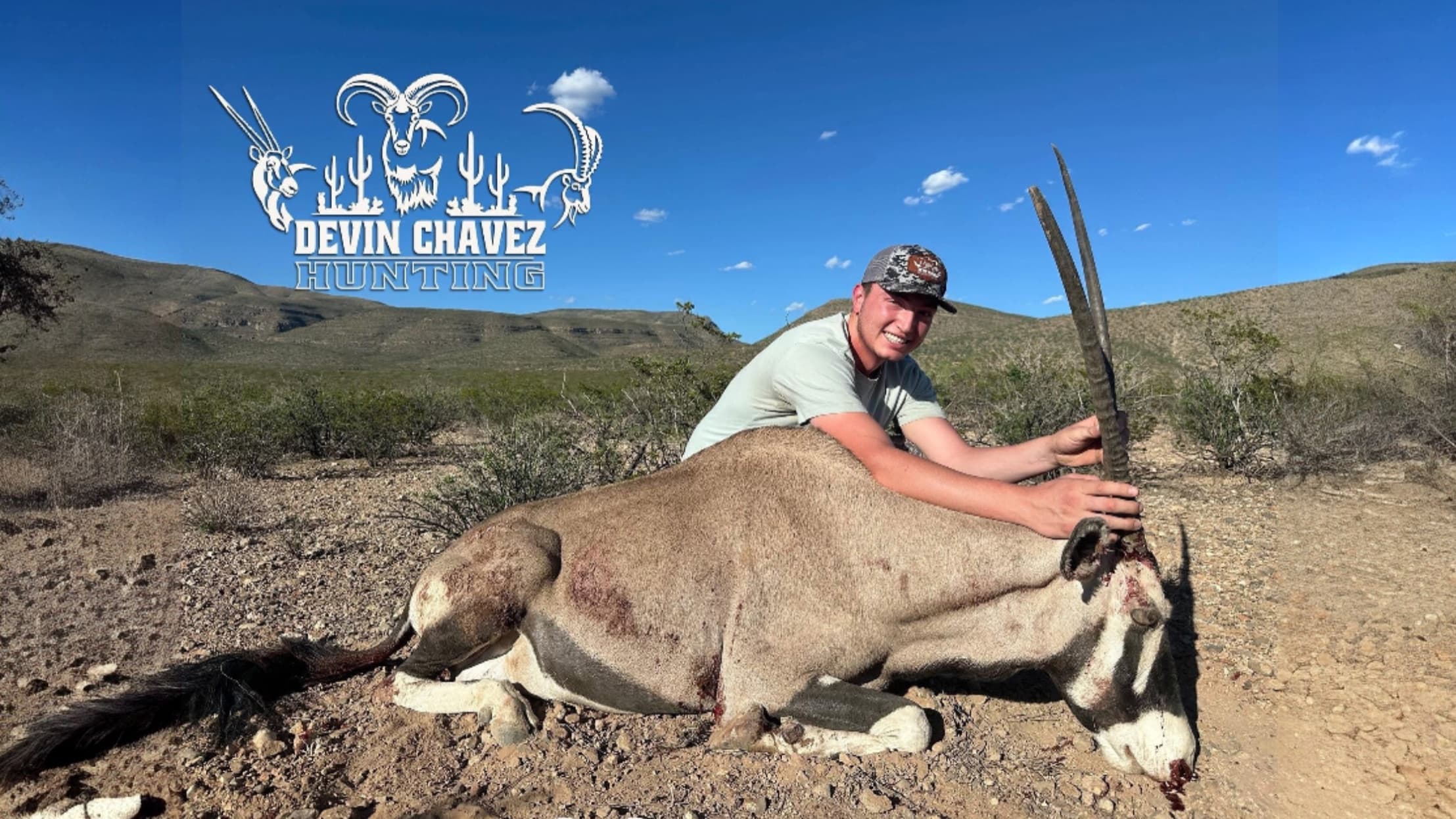 Man smiles, poses with an oryx he has hunted. The oryx lies on the ground with the man's hands on its long horns.